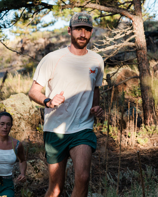 A man running outdoors on a wooded trail wearing a white short-sleeve performance t-shirt with a small red logo on the chest, dark green athletic shorts, white socks, and a camouflage baseball cap.